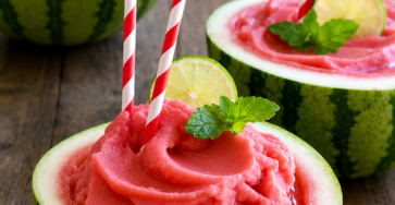 Two vibrant pink frozen watermelon slushies served inside hollowed-out watermelon bowls, topped with lime slices, fresh mint, and red striped straws.